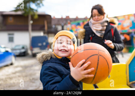 A five-year-old sweet boy stands in an amusement park square, holding a basketball ball, a blurry woman in the background. Stock Photo