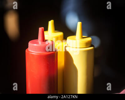 Three plastic bottles, red and yellow, used for fast food sauces such as ketchup and mustard, on display on a restaurant table with a black background Stock Photo