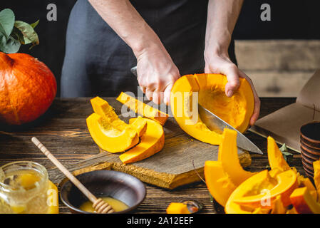 A woman cook cuts an orange pumpkin with a knife into slices for baking on a wooden table. Concept autumn food in a cozy dark kitchen with yellow flow Stock Photo