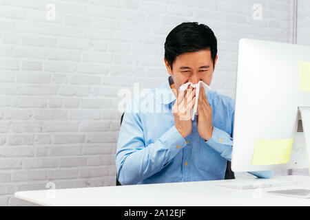Sick male entrepreneur sneezing with napkin at office with copy space. Asian young man with allergy and disease Stock Photo
