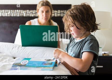 5 year old boy looking at book in hotel room as mom works on laptop Stock Photo