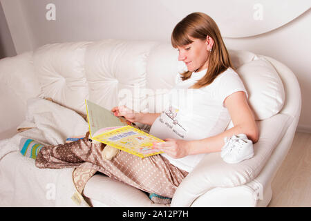 Zaporizhia, Ukraine - April 5, 2012: a young pregnant woman lies on a light sofa in her house, reads a book and smiles. Photo in light color. Stock Photo