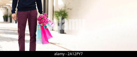 Pink flowers in man's hand and shopping bags on brick background. Birthday, Mother's, Women's, Wedding Day concept. Bunch of paper gift bags. Stock Photo