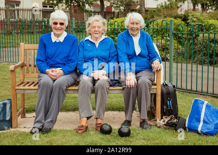 Three senior friends sitting on a bench at a bowling green, relaxing and smiling at the camera. Stock Photo