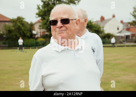 A portrait of a senior man smiling at a bowling green. He is looking away and wearing sunglasses. Stock Photo