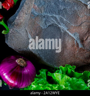 A wild multi-colored stone with a place for text lies on a black table next to fresh blue onions, yellow peppers, chili and lettuce Stock Photo