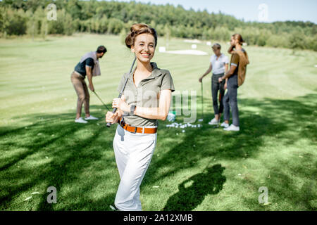 Portrait of an elegant young woman standing with golf putter and friends playing golf on the background Stock Photo