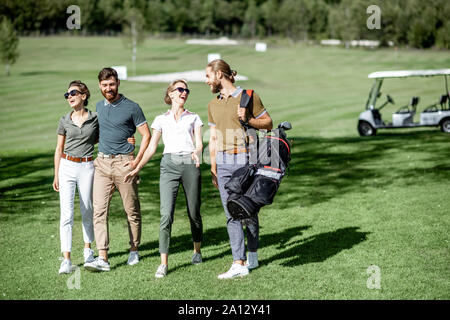 Young and elegant friends walking with golf equipment, hanging out together on the beautiful course with golf car on the background Stock Photo
