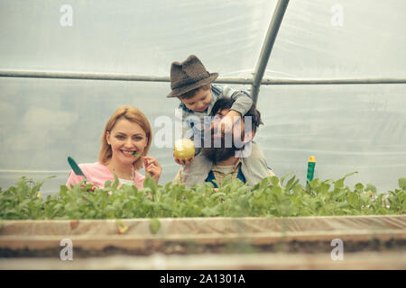 Happy family in greenhouse. Mum posing with green leaf in her mouth while kid is feeding dad with apple sitted on his shoulders. Bearded man playing w Stock Photo