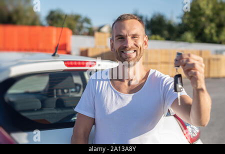 A male driver stands near a car with a key in his hand and smiles. Stock Photo