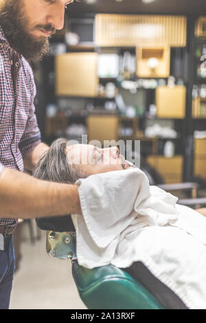 Barber covering client with towel before giving him a shave at barbershop. Stock Photo