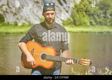 The guitarist plays the guitar, standing against the background of a river in the wild on a sunny summer day. A young bearded man in sunglasses, 23 ye Stock Photo