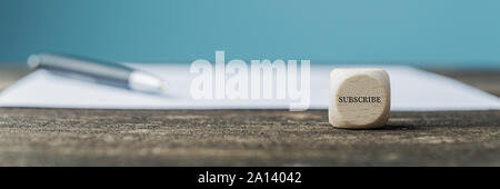 Wooden dice with the word Subscribe on it placed on wooden desk next to a blank paper or form and a pen. Stock Photo