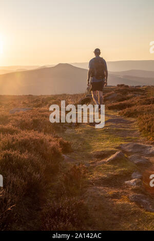 Man with a backpack walking on a moorland track as the golden sun is setting with a distant landscape of hills in the background. Stock Photo