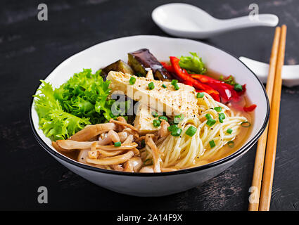 Asian vegan noodles soup with tofu cheese, shimeji mushrooms and fried eggplant in bowl on dark background.