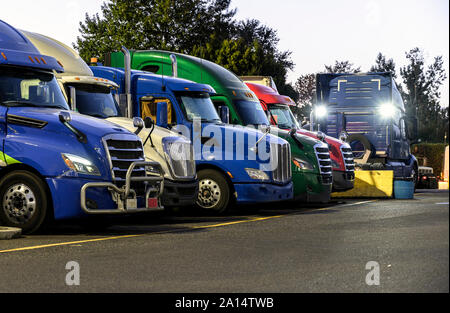 Different make and models big rigs semi trucks with semi trailers standing in row on truck stop parking lot for rest and comply with the movement acco Stock Photo