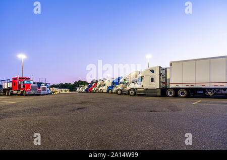 Different make and models big rigs semi trucks with semi trailers standing in row on truck stop parking lot for rest and comply with the movement acco Stock Photo