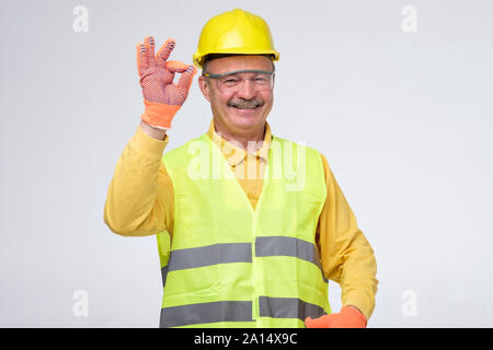 Senior hispanic construction worker in hard hat showing ok sign smiling on gray background Stock Photo