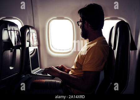 Adult bearded traveler man sitting inside the aircraft and flight to destination - computer with wifi internet connection on board service concept - t Stock Photo