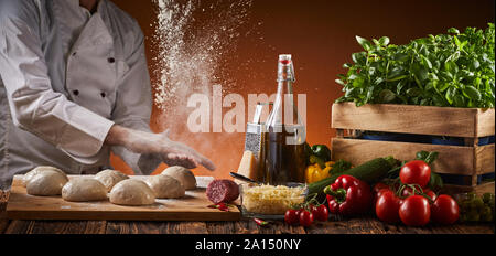 Freeze motion explosion of cooking flour as a chef preparing pizzas in a pizzeria claps his hands over portions of raw dough on a tray alongside fresh Stock Photo
