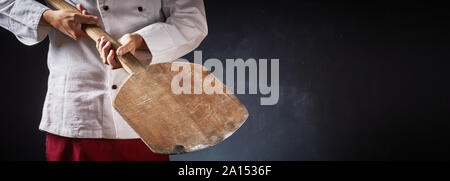 Chef holding an empty wooden pizza paddle in a close up on his hands in a panorama with dark background and copy space for food or product placement Stock Photo