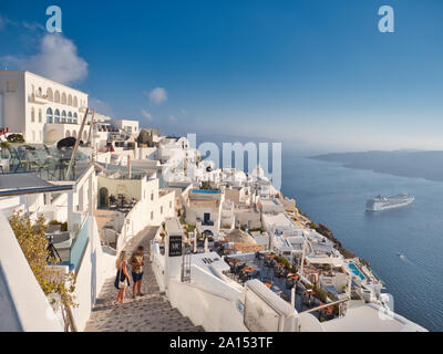 Picturesque view of famous white city of Fira on seashore with tourists and sailing ship in Santorini Greece on sunny day Stock Photo