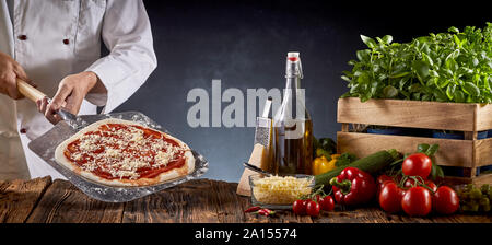 Chef making a traditional Margherita pizza holding a paddle with pie crust covered in tomato paste and cheese over a table with assorted fresh ingredi Stock Photo