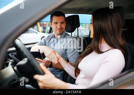 Male auto instructor takes exam in young woman. Shaking hands. Sitting in car. Brunette hold hand on steering wheel Stock Photo