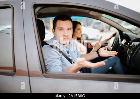 Male auto instructor takes exam in young woman. Wondered man look on camera. Confident female student driwing car and holding hands on steering wheel Stock Photo