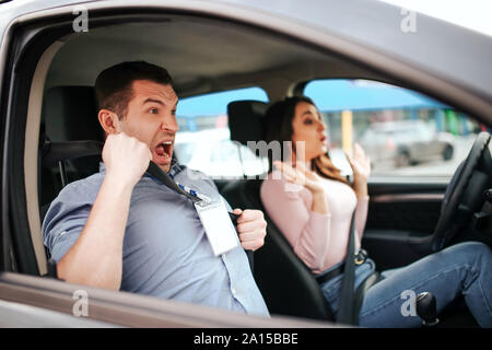 Male auto instructor takes exam in young woman. Scared guy screaming. Woman drives car careless. Hold hands not on steering wheel. Close to car crash Stock Photo