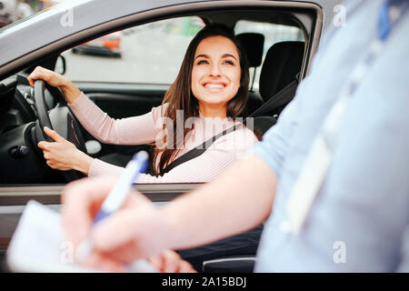 Male auto instructor takes exam in young woman. Cheerful attractive model look at guy and smile. Holding hands on steering wheel. Instructor writing m Stock Photo