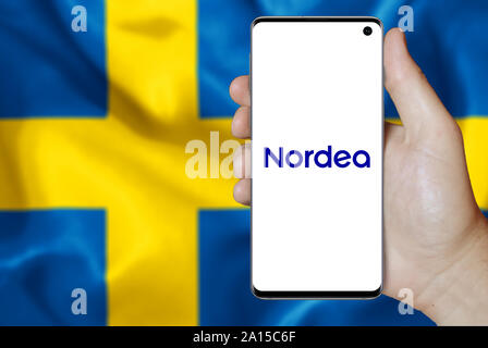 A man holds a smartphone displaying the logo of company Nordea listed on OMX Stockholm. Swedish flag in the background. Credit: PIXDUCE Stock Photo