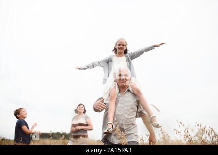 Happy girl sitting on her grandfather's shoulders in nature with brother and grandmother in the background Stock Photo