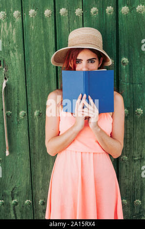Portrait of redheaded young woman standing in front of green wooden door covering mouth with a book Stock Photo