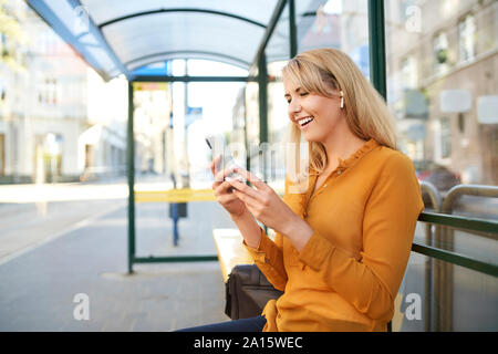 Happy young woman with wireless earphones using smartphone at bus stop