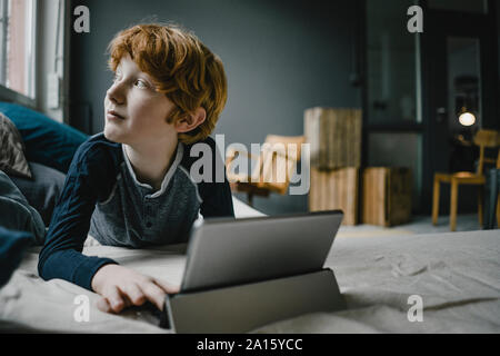 Redheaded boy lying on couch with digital tablet looking out of window