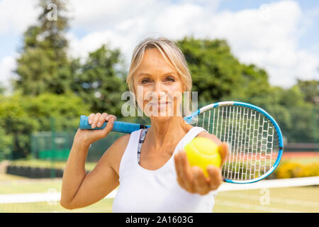 Portrait of smiling mature woman holding a tennis ball at tennis club Stock Photo