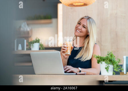 Portrait of smiling blond businesswoman with laptop and glass of orange juice in a coffee shop Stock Photo