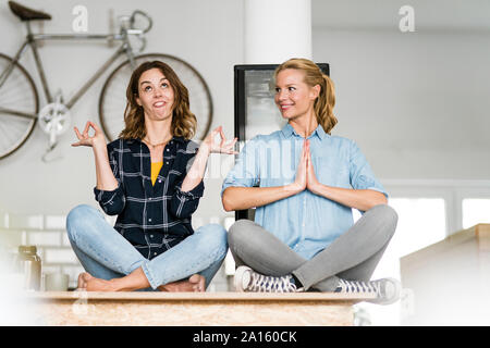 Two young women sitting cross-legged on the counter of their coffee shop, one meditating , other pulling funny faces Stock Photo