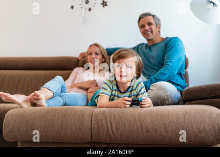 Boy lying on couch at home playing video game with parents watching Stock Photo