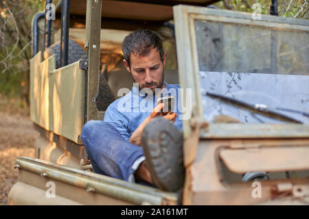 Man sitting in by off-road car, using smartphone Stock Photo