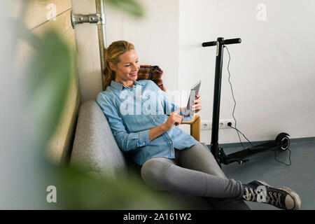 Young woman sitting on couch in a coffe shop, using digital tablet, e-scooter charging in background Stock Photo
