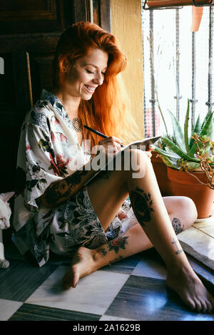 Red-haired tattooed woman at home writing in a notebook near the window Stock Photo