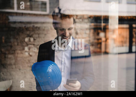 Smiling businessman with hard hat looking at cell phone Stock Photo