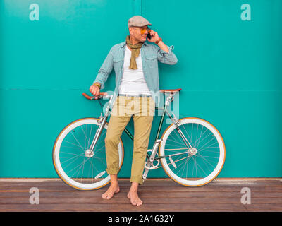 Barefoot man on the phone standing with Fixie bike in front of green wall Stock Photo