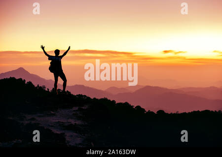 Hikers or travelers silhouette stands on mountain top against sunset with raised arms Stock Photo