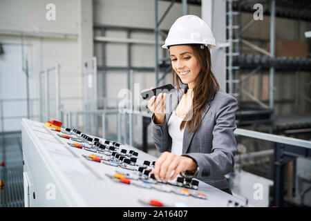 Woman wearing hard hat at control panel in a factory using cell phone Stock Photo