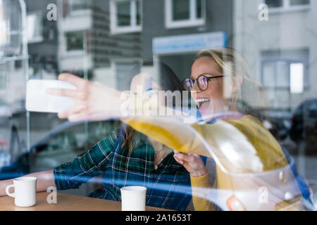 Two girlfriends sitting in a coffee shop, taking smartphone selfies Stock Photo
