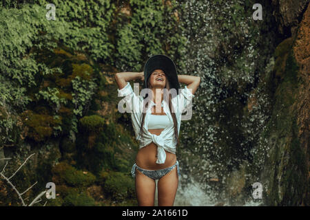 Happy young woman wearing a hat standing near a waterfall Stock Photo