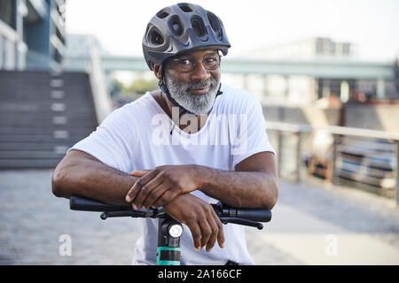 Portrait of smiling mature man wearing cycling helmet leaning on handlebar of Electric Scooter Stock Photo